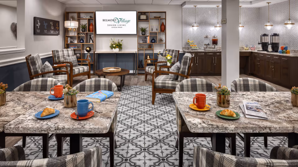 A cozy senior living common area with patterned tile flooring and granite tables set with colorful mugs, plates with croissants, a book, and a newspaper. The room features plaid upholstered chairs around the tables and wooden armchairs with plaid cushions arranged near a wall-mounted TV displaying the Belmont Village Senior Living Geneva Road logo. The back wall has dark wooden cabinets with a countertop holding coffee dispensers, fruit, and other refreshments under pendant lights.