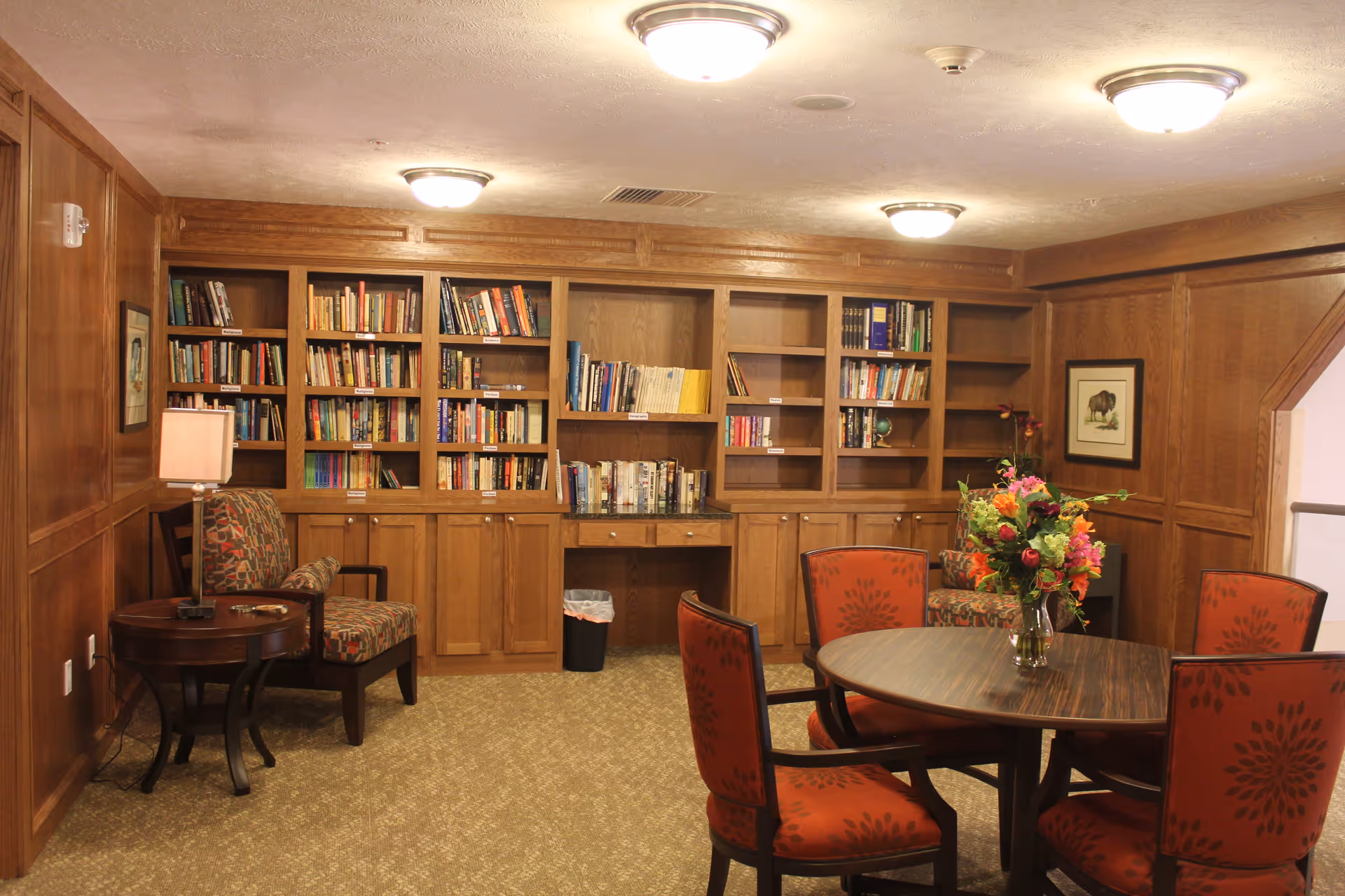 A cozy reading room with wooden bookshelves filled with books along the back wall. There is a round wooden table with four red upholstered chairs featuring a floral pattern. A vase with colorful flowers is placed on the table. To the left, there is a small round side table with a lamp and an upholstered armchair with a patterned cushion. The walls and ceiling are wood-paneled, and the room is softly lit by ceiling lights.