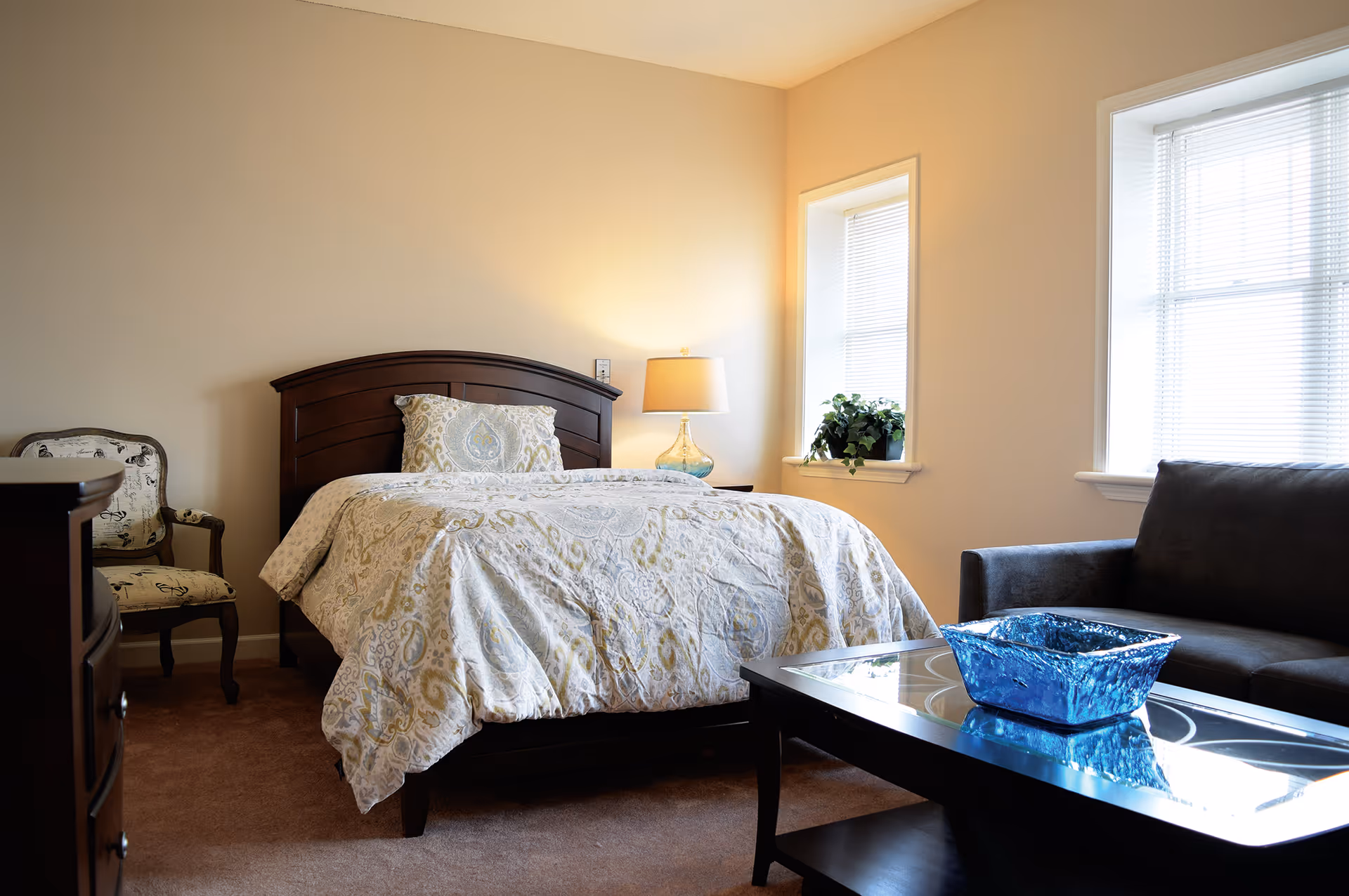 A cozy bedroom with a wooden bed frame and a patterned bedspread. There is a bedside table with a lamp, a cushioned armchair with a decorative pattern, a dark sofa, and a glass coffee table with a blue decorative bowl. Two windows with blinds allow natural light into the room.