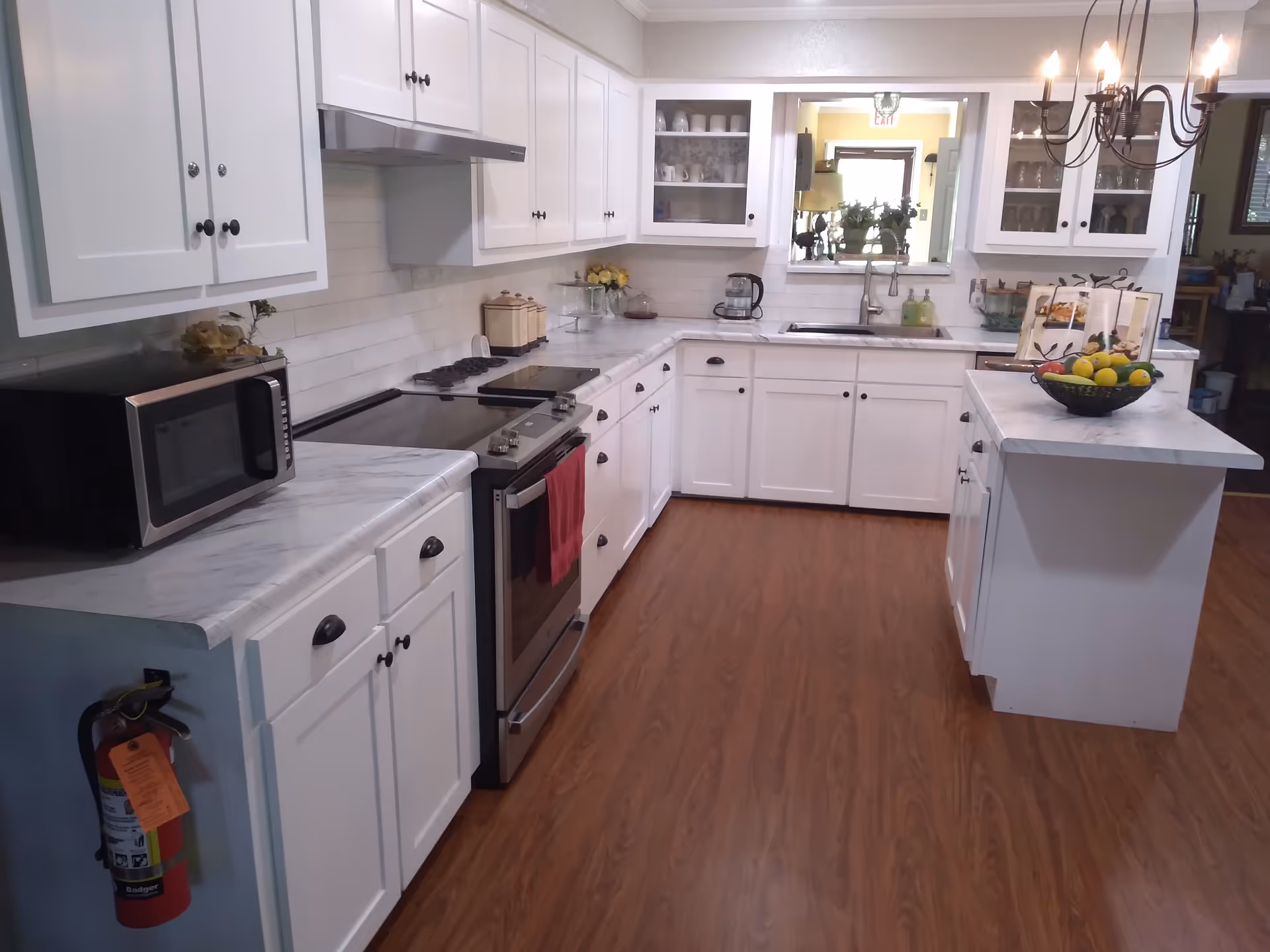 A clean and modern kitchen with white cabinets, a marble countertop, a stove with a red towel hanging on the handle, a microwave, a sink under a window, and a small island with a bowl of fruit. The floor is wooden, and a chandelier with lit bulbs hangs above the island.