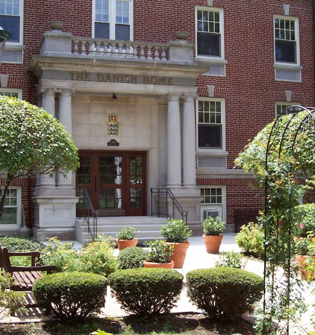 Entrance of The Danish Home building with a brick exterior, stone columns, and a small garden area with trimmed bushes and potted plants in front.