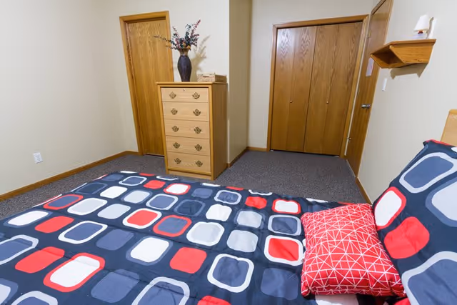A bedroom with a bed covered in a colorful geometric patterned bedspread in shades of blue, red, white, and gray. There is a red pillow with a white geometric pattern on the bed. The room has beige walls, a wooden dresser with a vase of flowers on top, a wooden door, and a wooden closet with double doors.