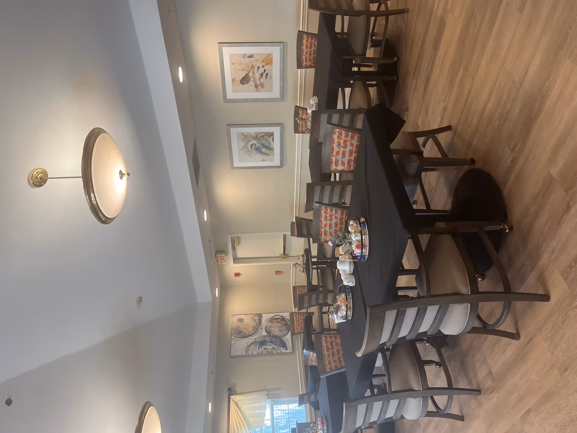 A dining room with several tables covered in black tablecloths and surrounded by wooden chairs with cushioned seats and patterned backs. Each table has a centerpiece with cups, plates, and small decorative items. The room has light-colored walls adorned with framed bird artwork and a window with a valance. The floor is wood, and ceiling lights provide illumination.