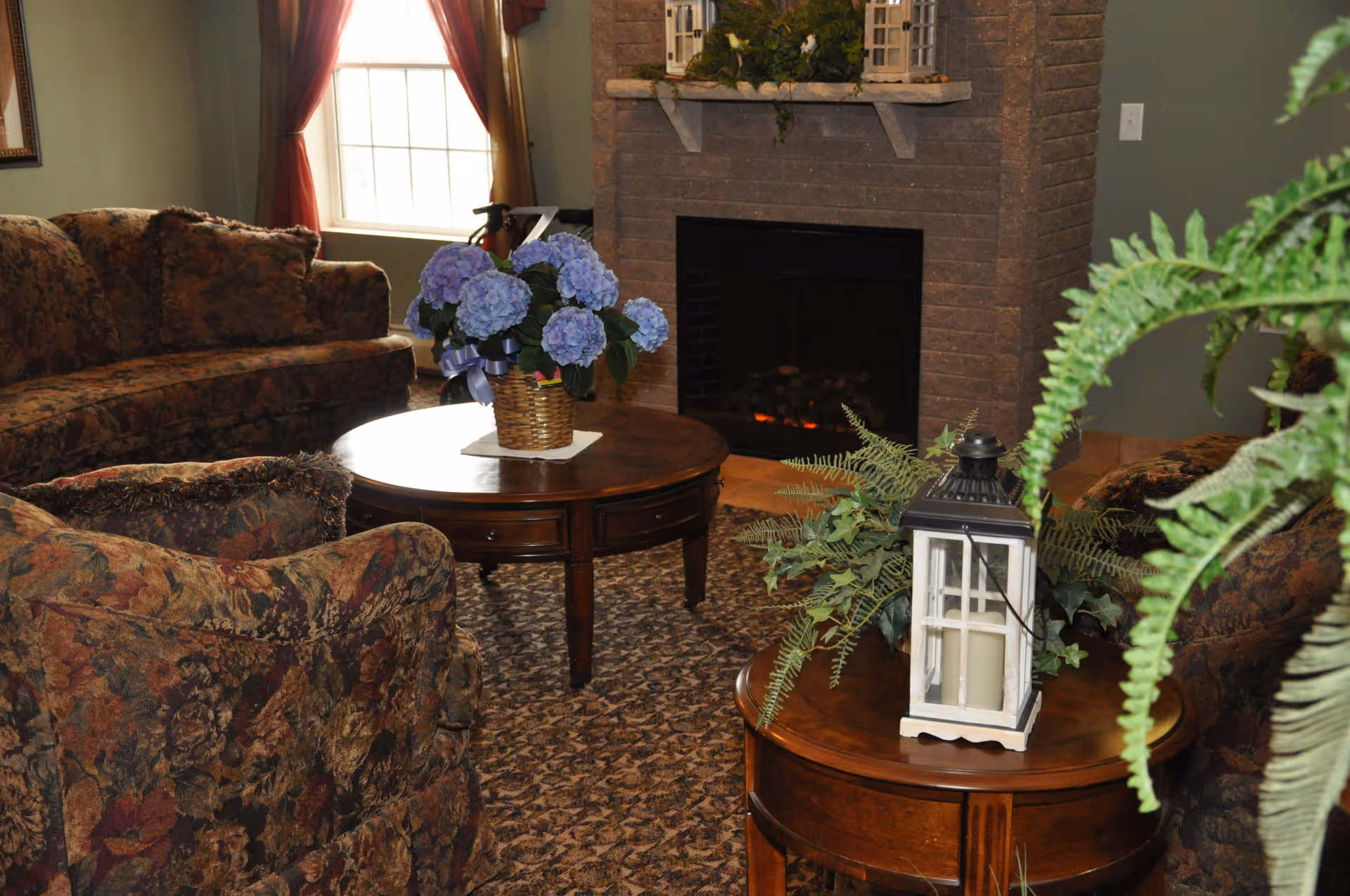 A cozy living room with floral patterned sofas and armchairs arranged around two wooden coffee tables. One table has a basket of blue hydrangeas, and the other has a white lantern and green plants. A brick fireplace is in the background, with decorative items on the mantel. A window with red curtains allows natural light into the room.