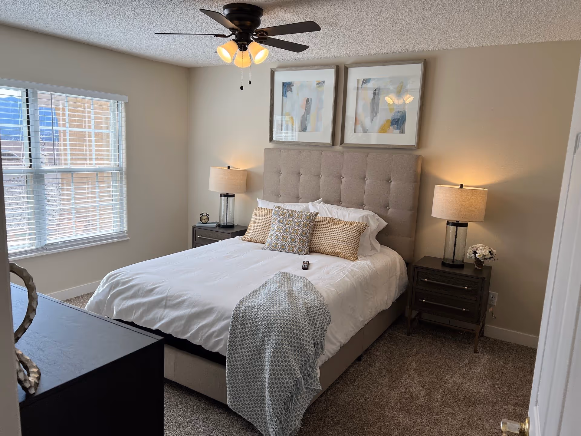 A neatly made bed with a beige tufted headboard in a bedroom. The bed has white bedding, three decorative pillows, and a light blue throw blanket draped over the foot. There are two matching dark wood nightstands on either side of the bed, each with a lamp and one with a small flower arrangement. Above the headboard are two abstract framed artworks. A ceiling fan with lights is mounted on the ceiling, and a window with white blinds is on the left wall letting in natural light. The room has beige walls and carpeted floor.