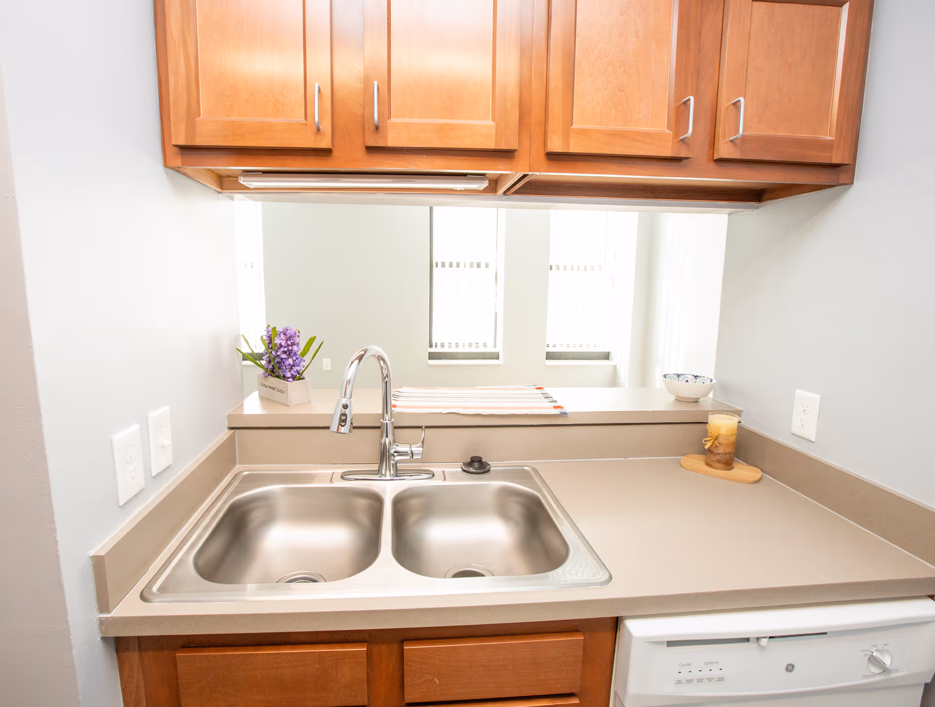 A clean kitchen countertop with a double stainless steel sink and a modern faucet. Above the sink are wooden cabinets. On the countertop, there is a small potted plant with purple flowers, a striped cloth, a decorative bowl, and a candle on a wooden tray. A dishwasher is visible below the counter.