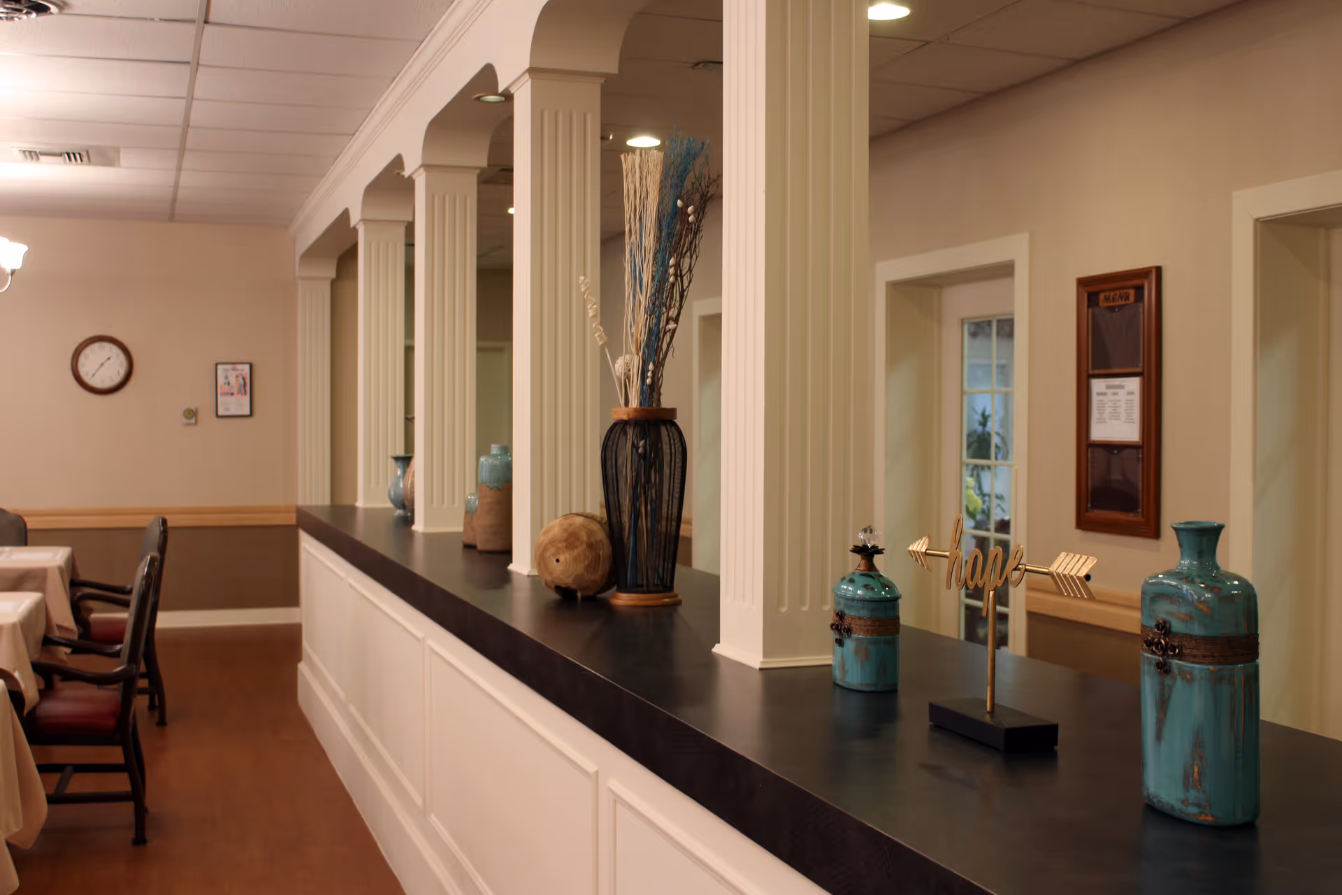 Interior view of a senior living facility dining area with a long counter decorated with vases and a decorative 'hope' sign. There are columns along the counter and tables with chairs in the background. The walls are beige with a clock and framed items hanging.