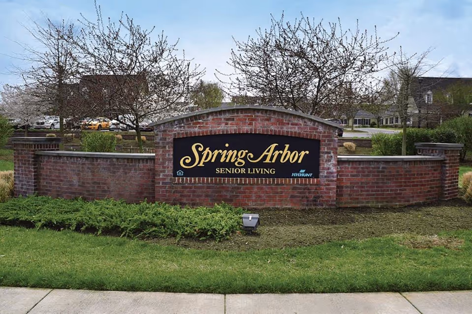A brick sign for Spring Arbor Senior Living is displayed outdoors with trees and buildings in the background. The sign is set in a landscaped area with green grass and shrubs.