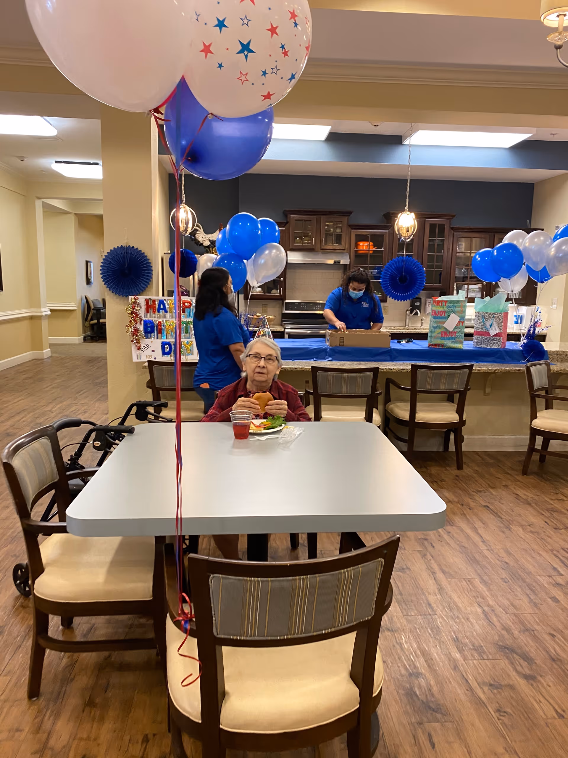 An elderly woman sitting at a table eating a sandwich and drinking a red beverage in a decorated room with blue, white, and red balloons. Two staff members in blue shirts are in the background near a kitchen counter with gift bags and a Happy Birthday sign.