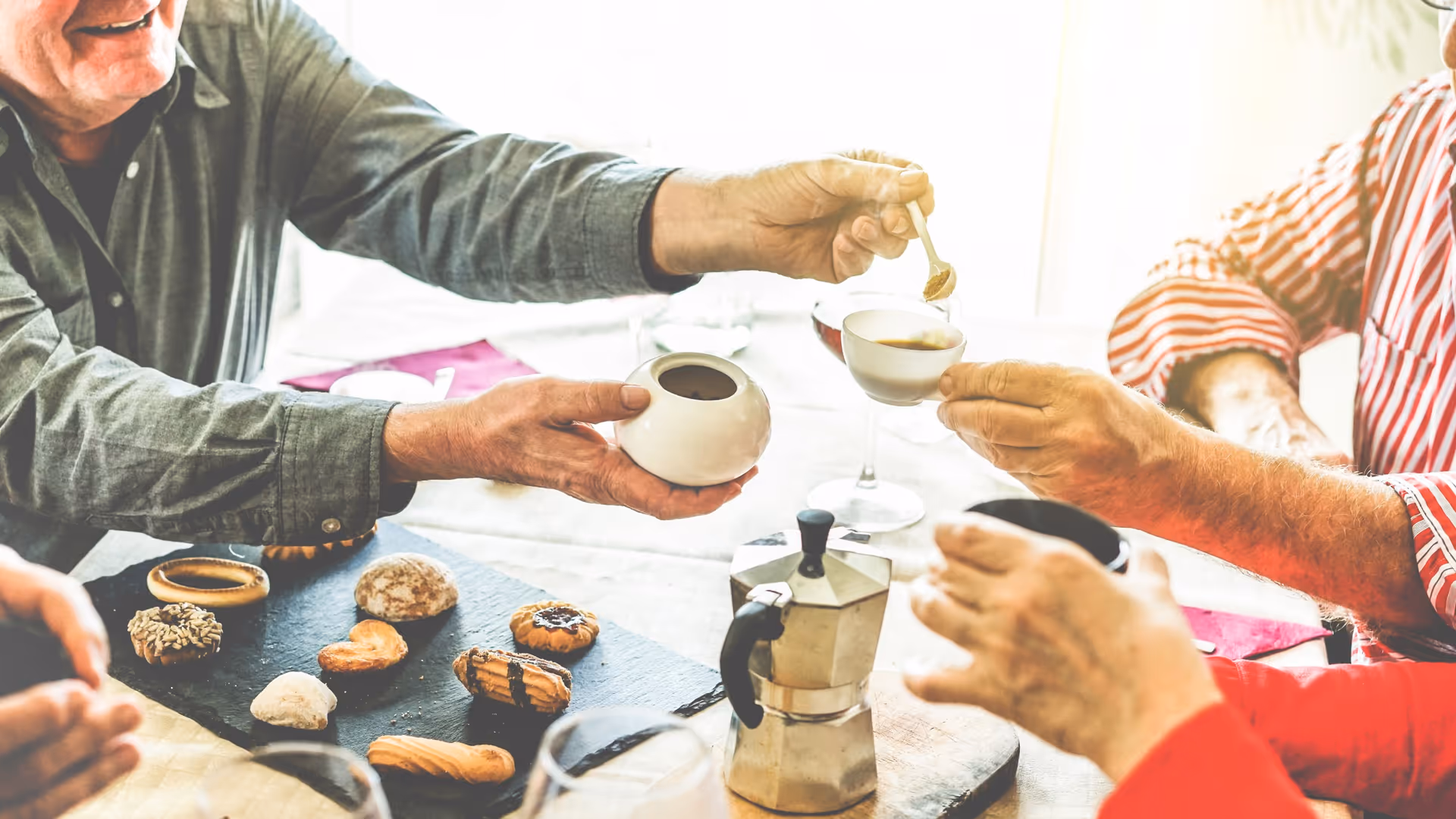 Three elderly people sitting around a table sharing coffee and cookies. One person is holding a small white sugar bowl and spoon, adding sugar to a cup held by another person. Various cookies and a stovetop coffee maker are on the table.