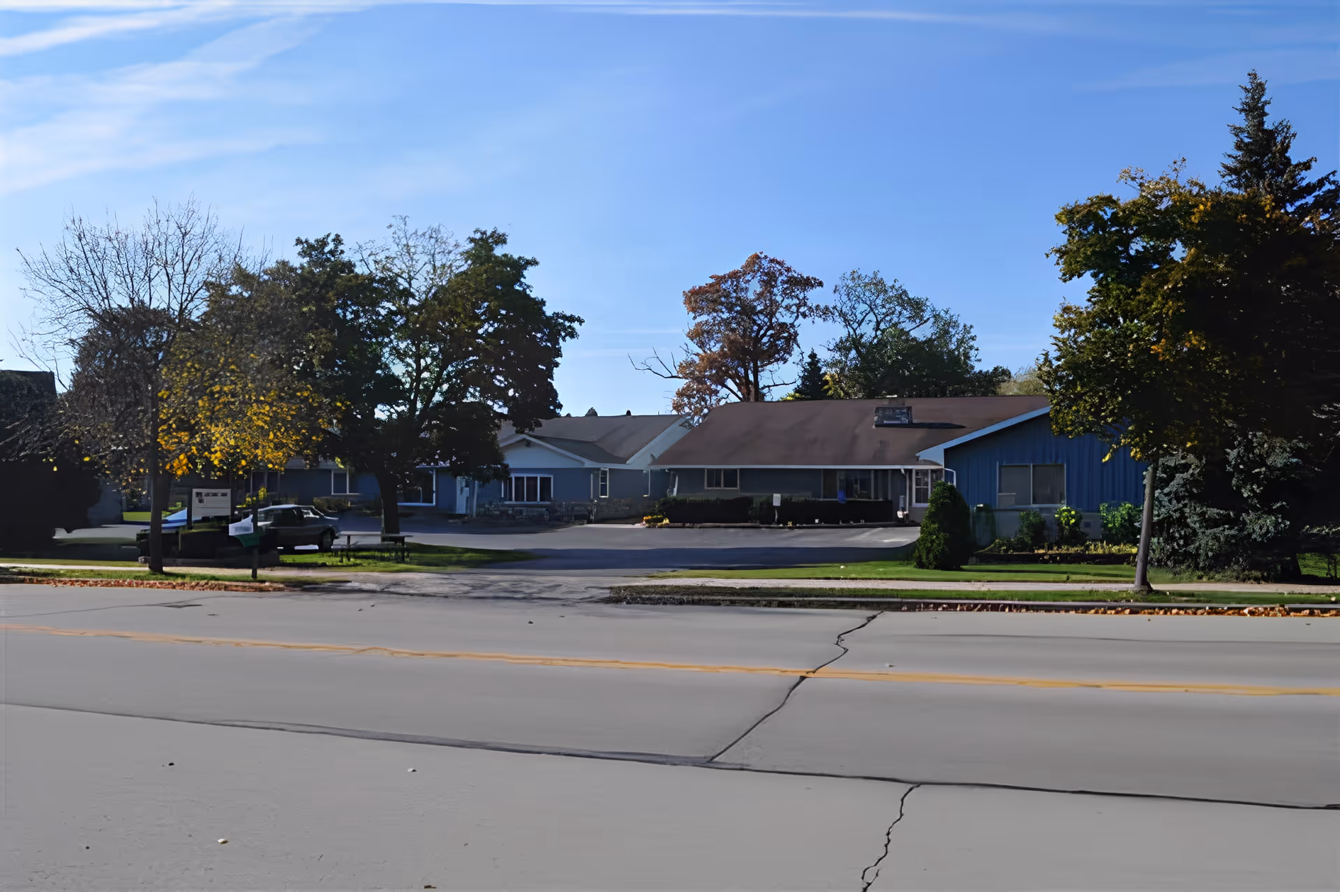 Exterior view of Autumn Oaks Assisted Living facility showing a single-story building with a brown roof and blue siding, surrounded by trees with some autumn foliage, and a paved road in the foreground.