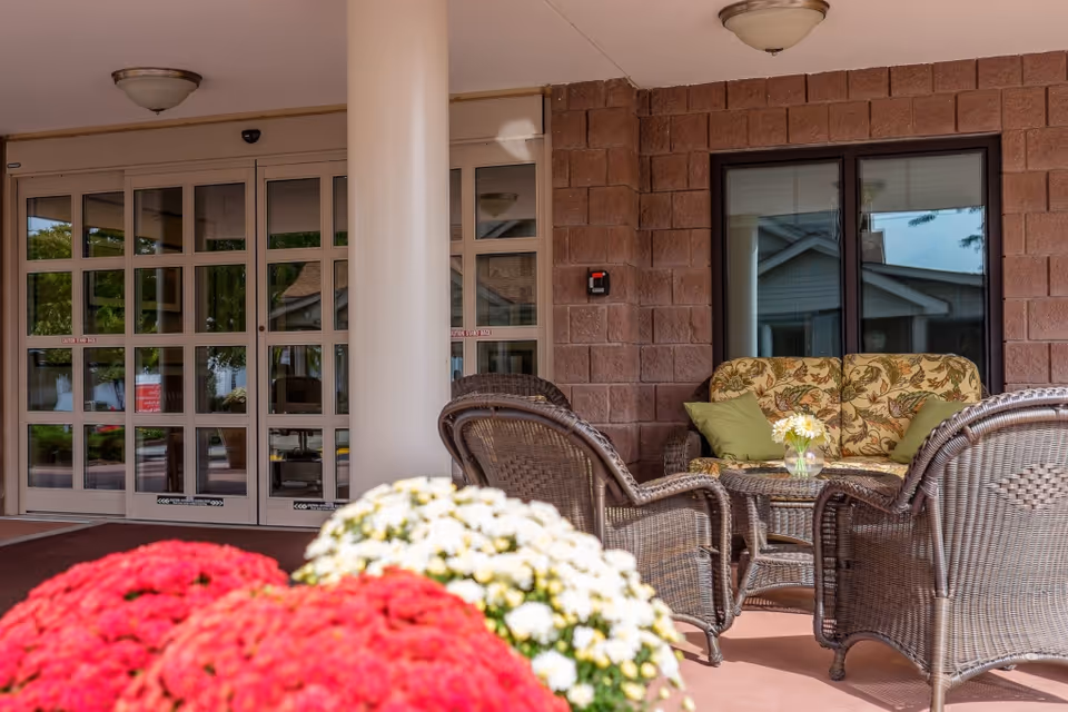 Outdoor seating area at the entrance of Beechwood at Eddy Memorial featuring wicker chairs and a cushioned loveseat with floral patterned cushions and green pillows, a small round table with a vase of flowers, red and white flowers in the foreground, and glass double doors leading inside.