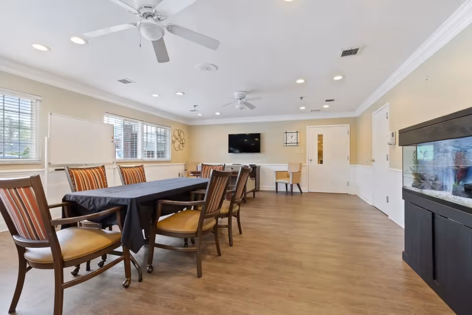 A bright communal activity room with a long table covered by a dark cloth surrounded by chairs, a wall-mounted TV, whiteboard, and an aquarium.