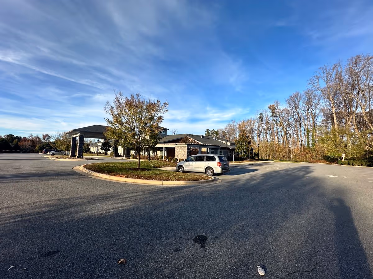 Front exterior of a single-story senior living building with a covered entrance, parking lot, trees, and a parked van under a blue sky.
