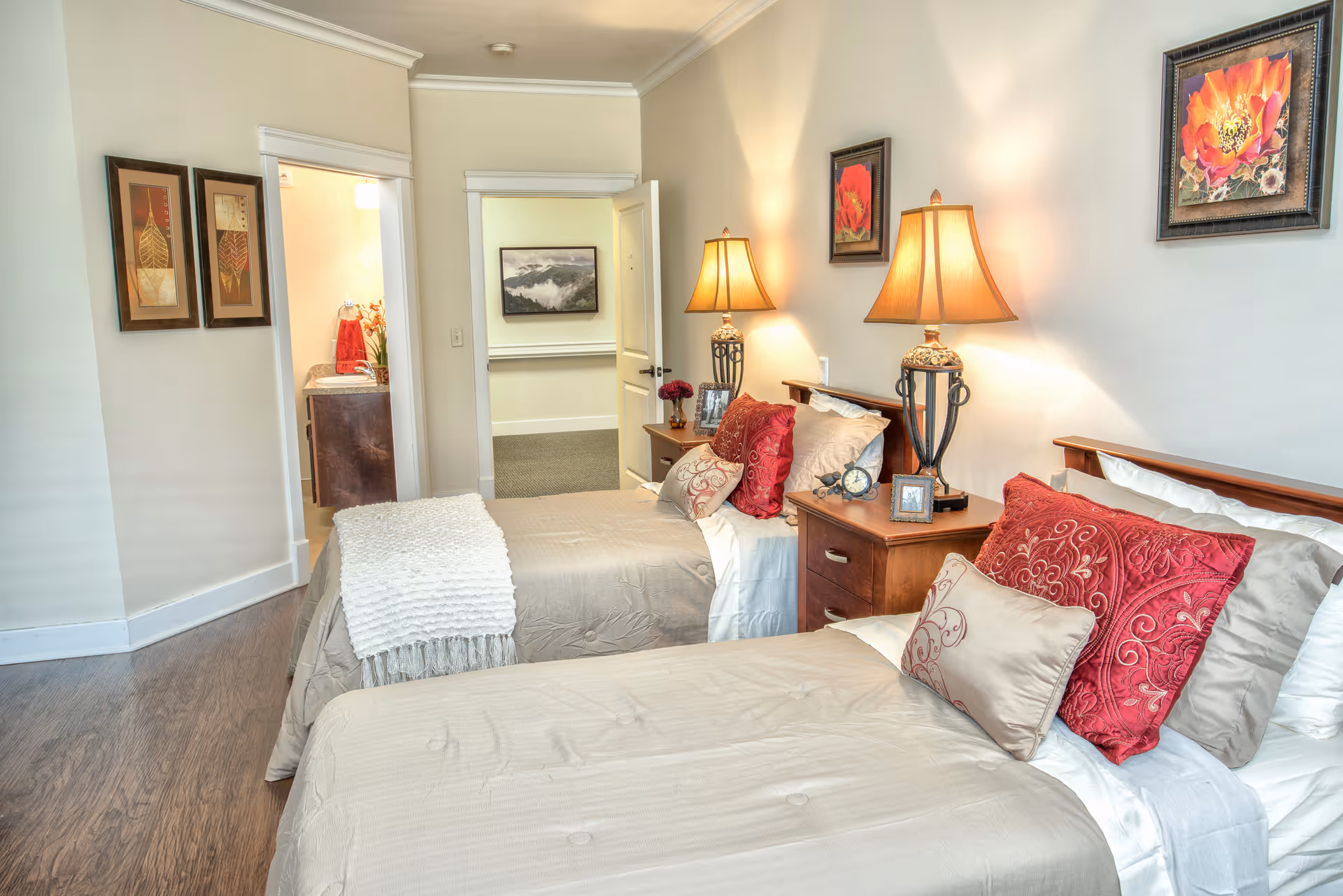A bedroom with two single beds side by side, each with beige bedding and decorative pillows in red and beige. Between the beds is a wooden nightstand with two matching table lamps, a clock, and framed photos. The walls are light-colored with framed floral artwork. A doorway leads to a bathroom and another room with a carpeted floor.