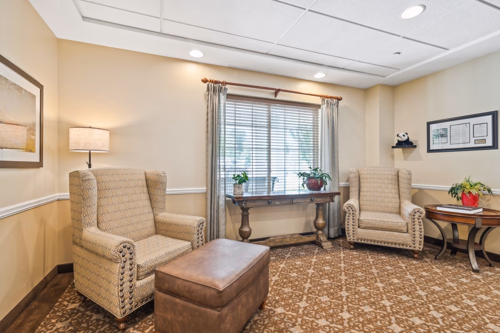 Comfortable sitting area with two upholstered armchairs, a leather ottoman, side tables, and a window with blinds.