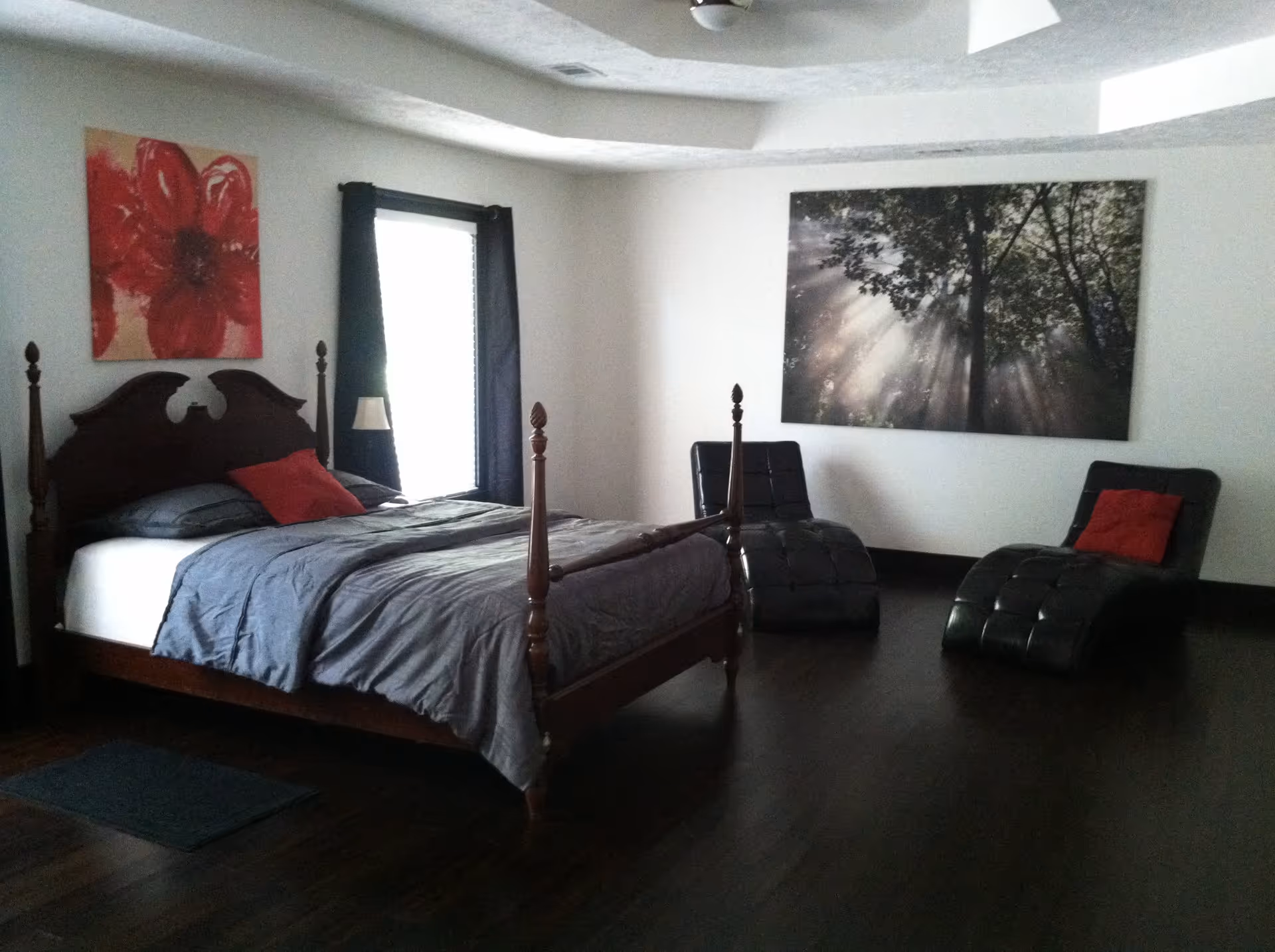 A bedroom with a dark wooden four-poster bed made with gray bedding and a red accent pillow. There are two black chaise lounge chairs with red pillows positioned near a wall. The room has dark hardwood floors, a window with black curtains, and two large pieces of wall art: one with a red flower and another depicting sunlight filtering through trees.