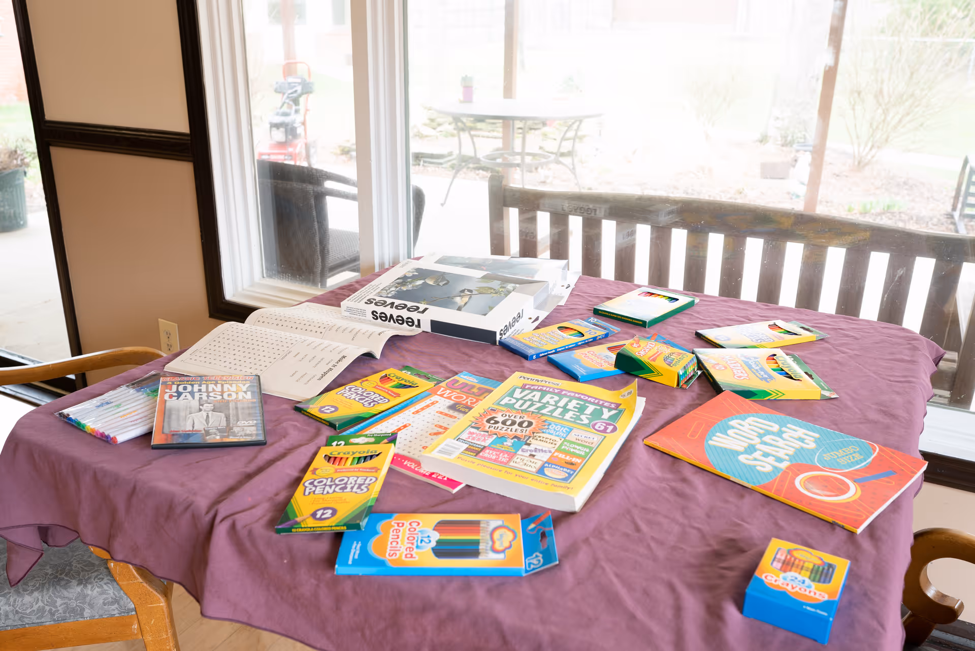 A table covered with a purple tablecloth is set up near large windows overlooking an outdoor patio. On the table are various activity books including word search and puzzle books, several packs of colored pencils and crayons, a DVD case, and some papers. Chairs are positioned around the table.