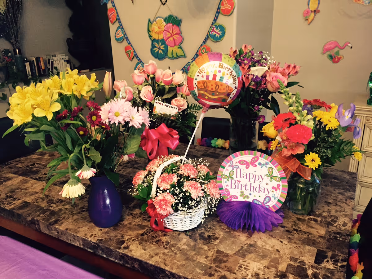 A marble table decorated with multiple colorful flower arrangements in vases and a basket, along with birthday decorations including a balloon and a sign that says 'Happy Birthday'. The background shows a wall with a floral banner and some flamingo decorations.