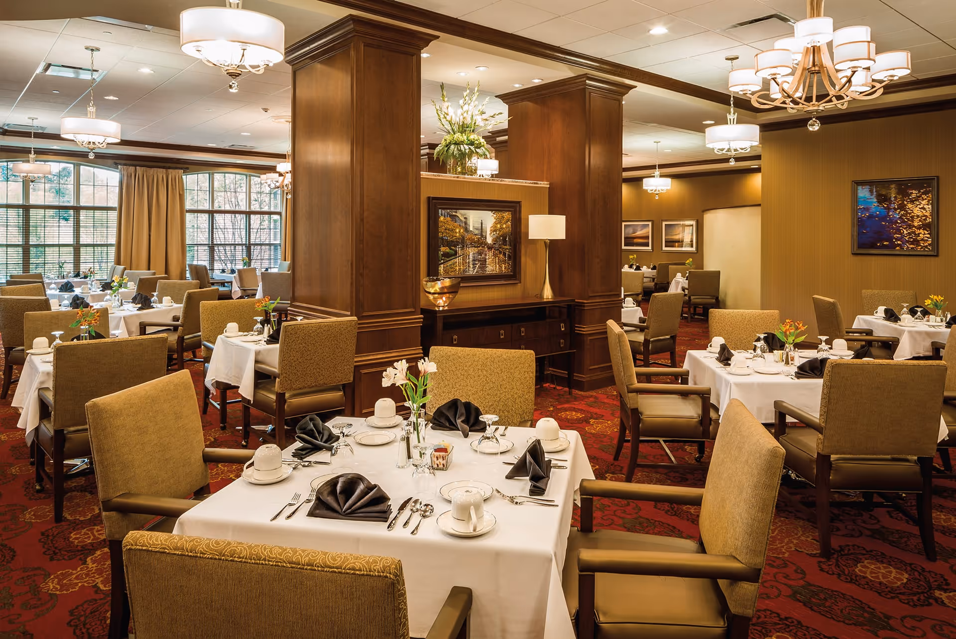 A well-lit dining room with multiple tables covered in white tablecloths, each set with plates, cups, silverware, and black folded napkins. The room features brown upholstered chairs, wooden columns, framed artwork on the walls, and large windows with beige curtains allowing natural light to enter.