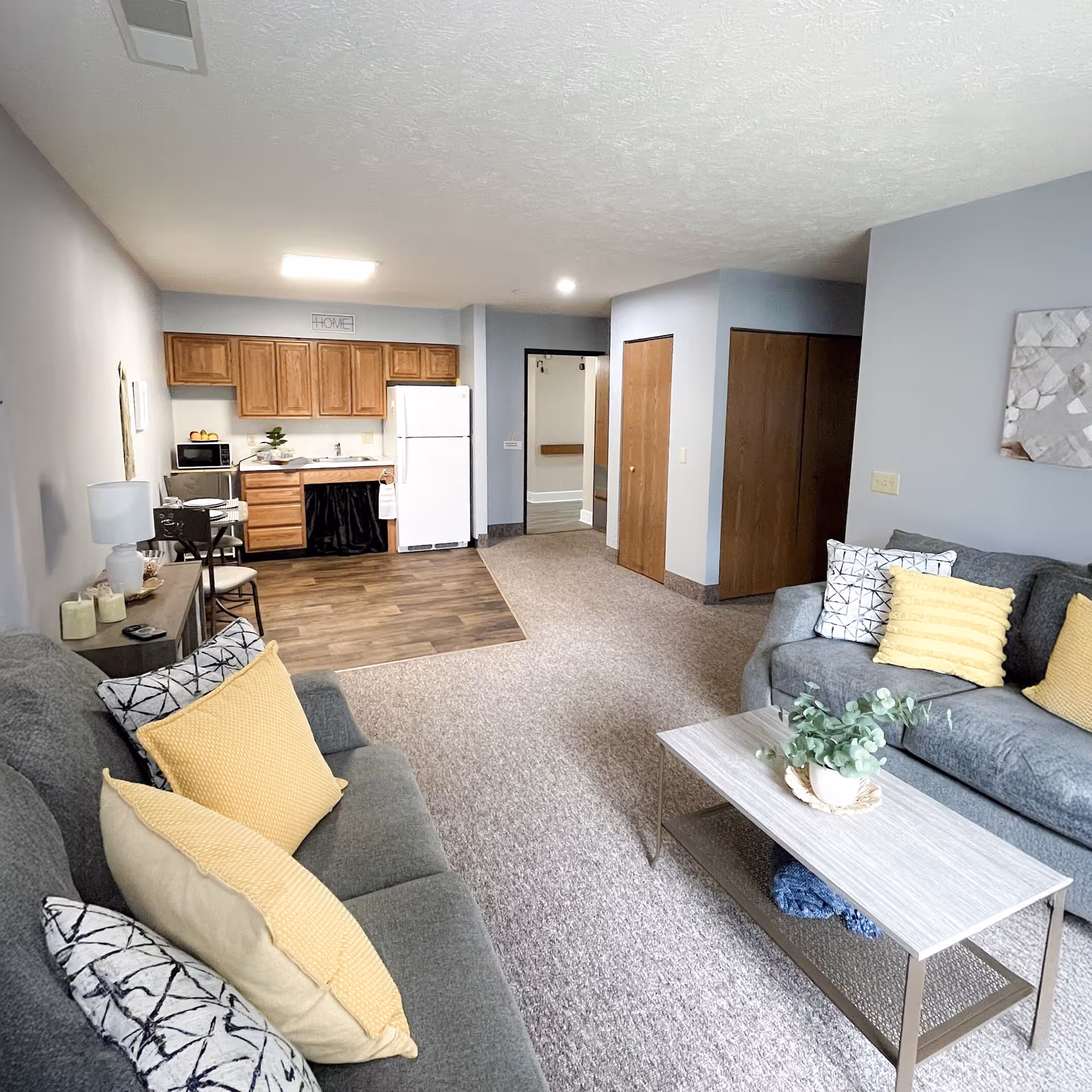 A cozy senior living apartment interior featuring a small kitchen area with wooden cabinets, a white refrigerator, microwave, and sink. The living area has two gray sofas adorned with yellow and patterned pillows, a light-colored coffee table with a plant, and a side table with a lamp and candles. The walls are painted light gray, and there is a carpeted floor with a section of wood flooring in the kitchen area. A hallway and closets are visible in the background.