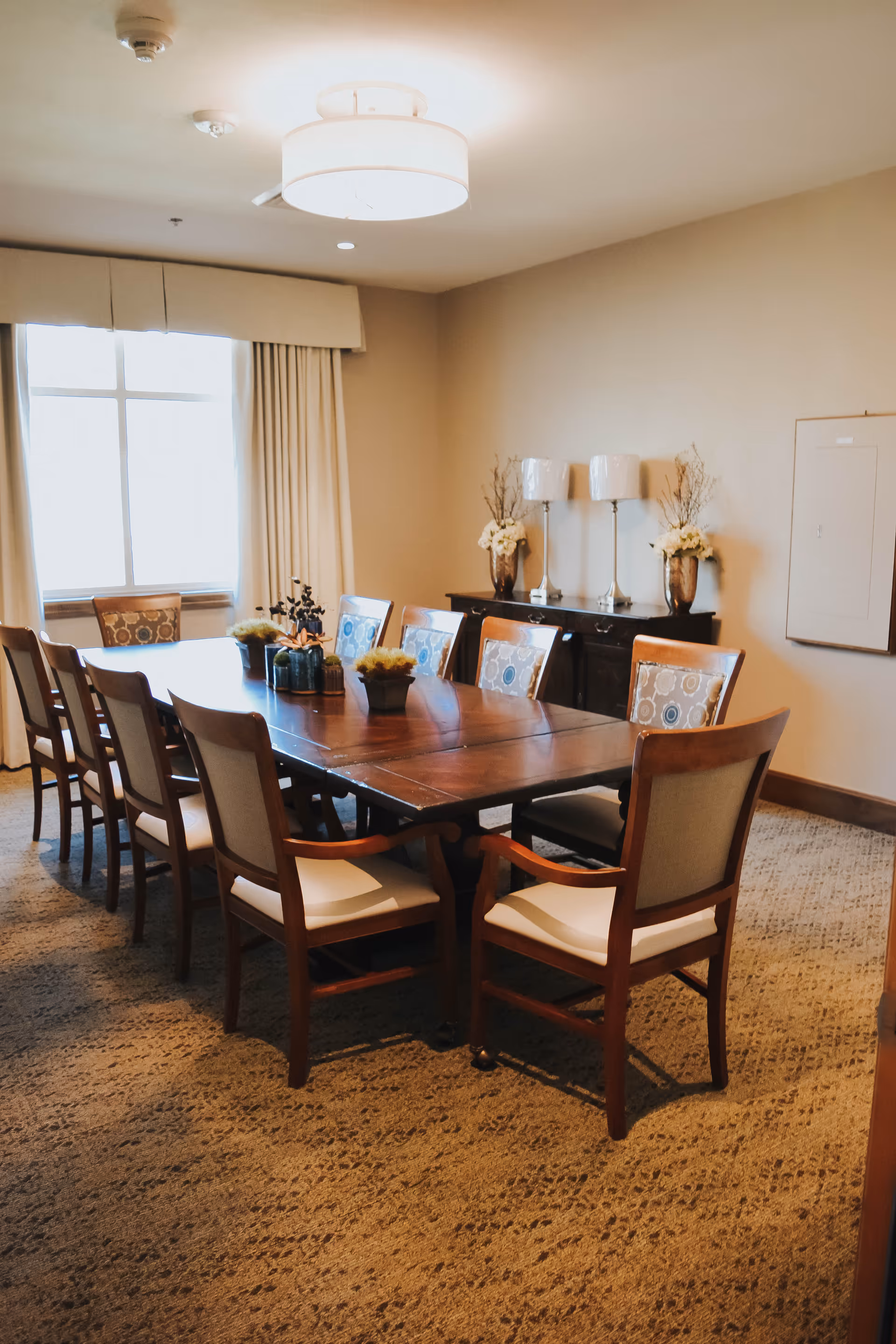 A well-lit dining room with a long wooden table surrounded by ten cushioned chairs. The room has beige walls and carpeted floors. A window with curtains allows natural light to enter. Against one wall is a dark wooden sideboard with two table lamps and decorative vases with flowers.