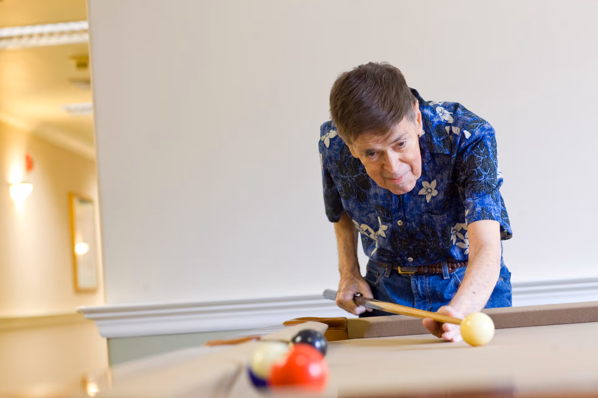An elderly man wearing a blue floral shirt is playing pool indoors, aiming to strike the cue ball with a pool cue on a billiards table.