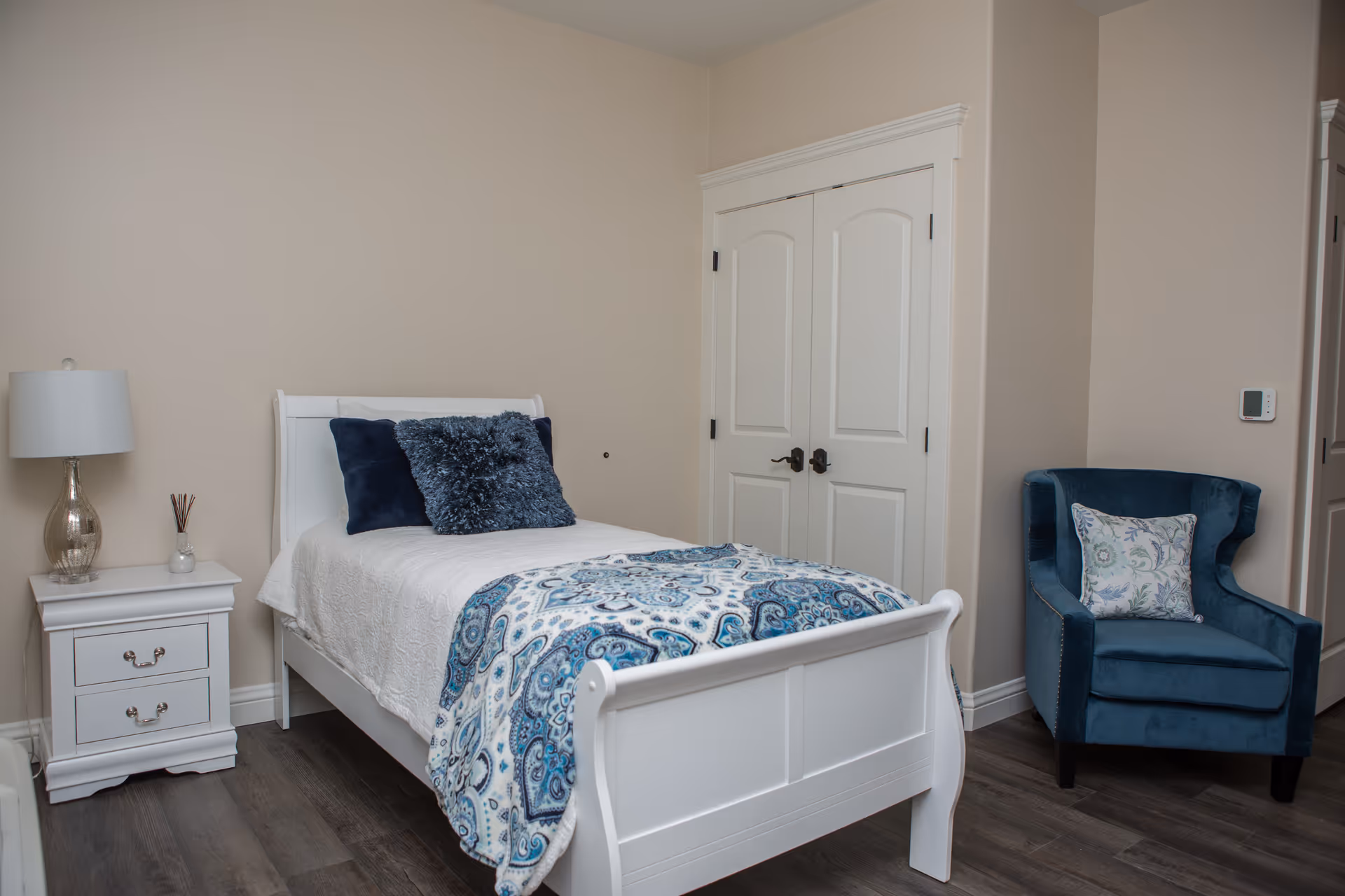 White bedroom with a white bed, nightstand, lamp, and blue accent chair.