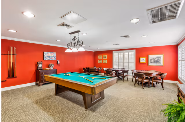 A recreational room with a pool table in the center, surrounded by several wooden tables and chairs. The walls are painted red and decorated with framed pictures. There is a rack with pool cues on one wall and a small cabinet with decorative items. The room has carpeted flooring and ceiling lights.