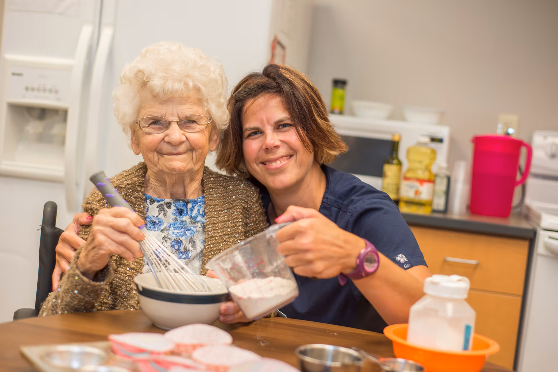 An elderly woman and a younger woman smiling together in a kitchen setting. The elderly woman is holding a whisk and a bowl, while the younger woman is holding a measuring cup with batter. They appear to be baking or cooking together.