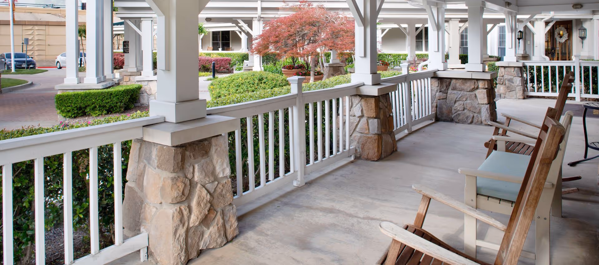 Covered outdoor porch area with wooden rocking chairs and a bench with cushions, surrounded by white railings supported by stone pillars. In the background, there are landscaped bushes, a small tree with red leaves, and parts of the building exterior with windows and doors.