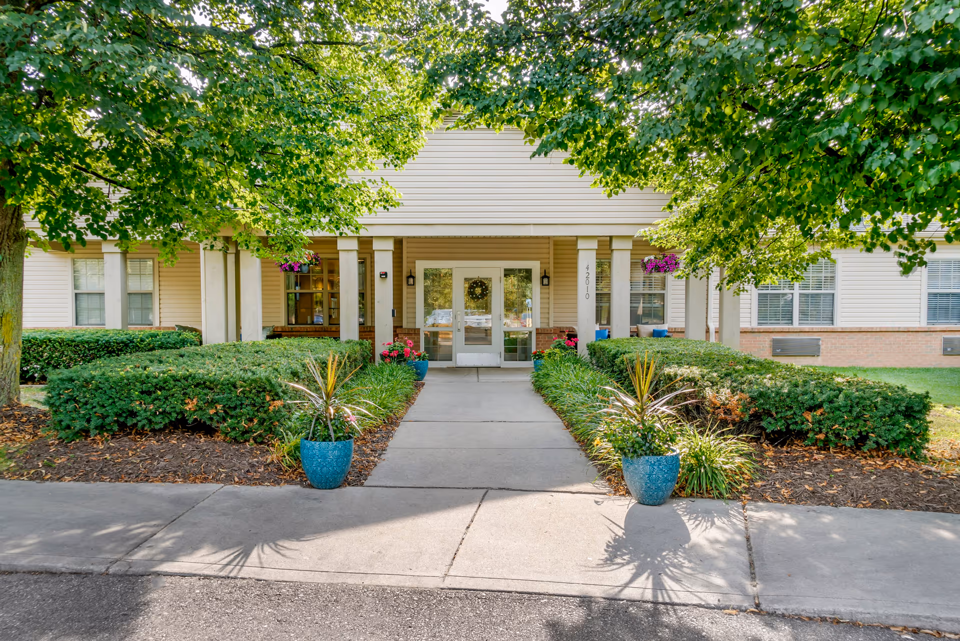 Entrance to a senior living facility with a concrete walkway flanked by green shrubs and two blue planters with plants. The building has beige siding, white columns, and a glass door with a wreath. Trees with green leaves frame the entrance.