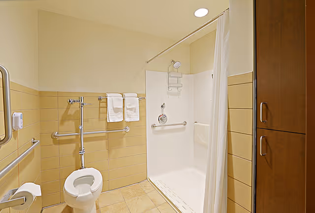 Accessible bathroom with a toilet, multiple grab bars, towel rack, walk-in shower with curtain, and a wooden storage cabinet.