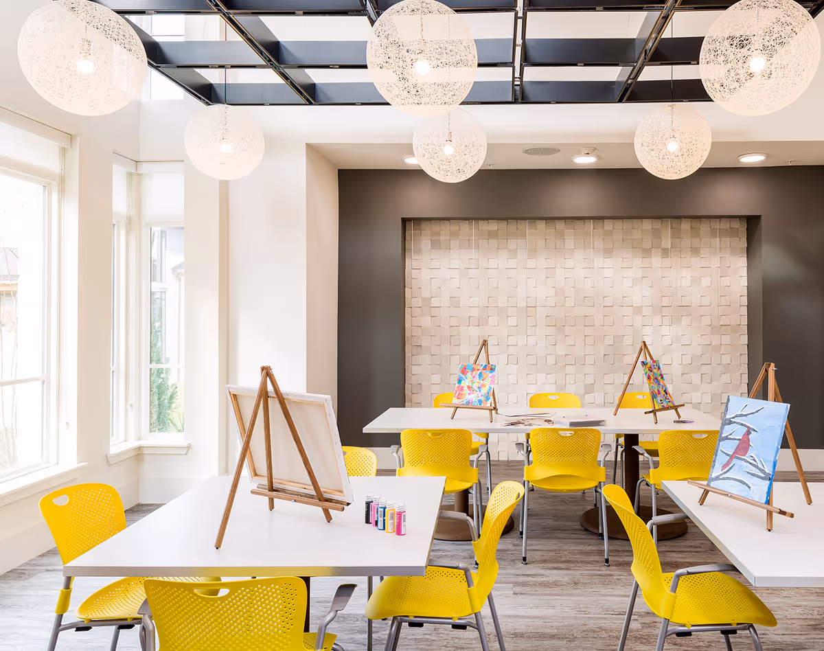 Bright art room with large windows letting in natural light, featuring white tables and yellow chairs. Several easels hold colorful paintings, and paint bottles are visible on one table. The ceiling has modern spherical pendant lights, and the back wall has a textured design.