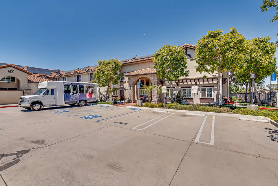 Exterior view of Pacifica Senior Living South Coast building with a shuttle bus parked in the handicap parking area. The building has a tiled roof, stone arch entrance, and several trees in front. The parking lot is mostly empty under a clear blue sky.