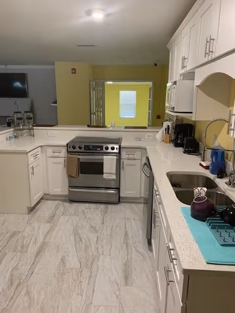 Modern kitchen with white cabinets and drawers, a stainless steel oven and stove, a microwave, a coffee maker, a sink with a faucet, and various kitchen items on the countertops. The floor has light-colored tiles and there is a window opening to another room with yellow walls.