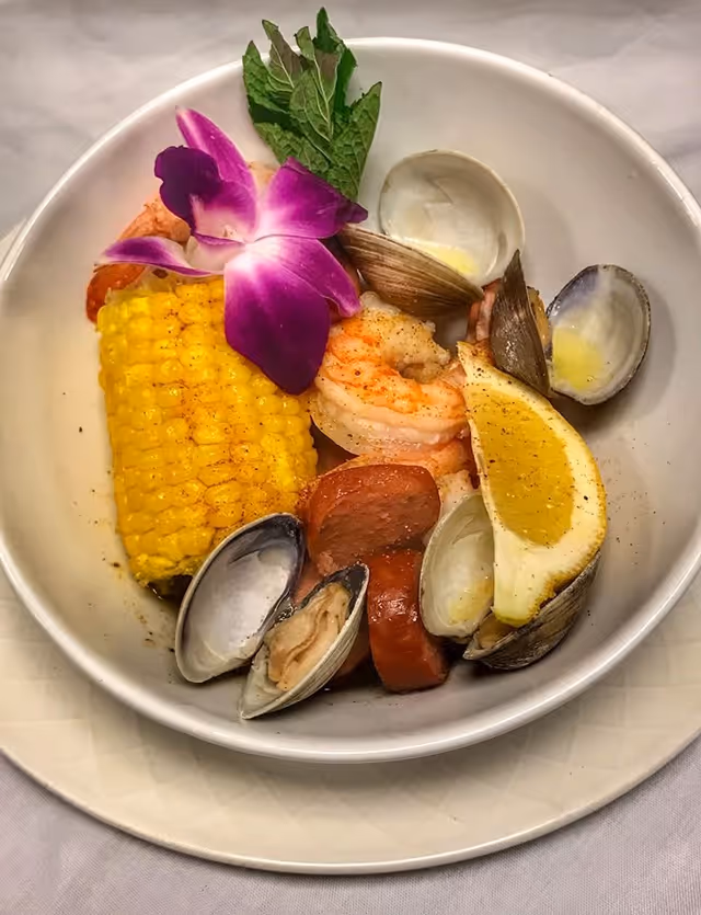 A bowl of seafood boil containing shrimp, clams, slices of sausage, a piece of corn on the cob, a lemon wedge, garnished with a purple edible flower and a sprig of mint.