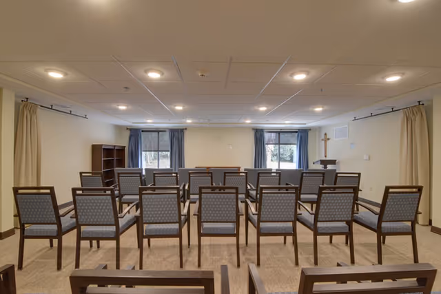 A small chapel/meeting room with rows of chairs facing a podium and a wall-mounted cross under two windows.