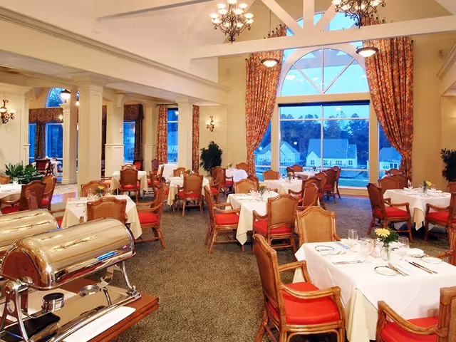 A spacious dining room with multiple tables covered in white tablecloths, set with plates, glasses, and silverware. The room features wooden chairs with red cushions, large windows with floral curtains, and chandeliers hanging from a high ceiling with exposed beams. There are also two silver buffet warmers on a wooden table in the foreground.