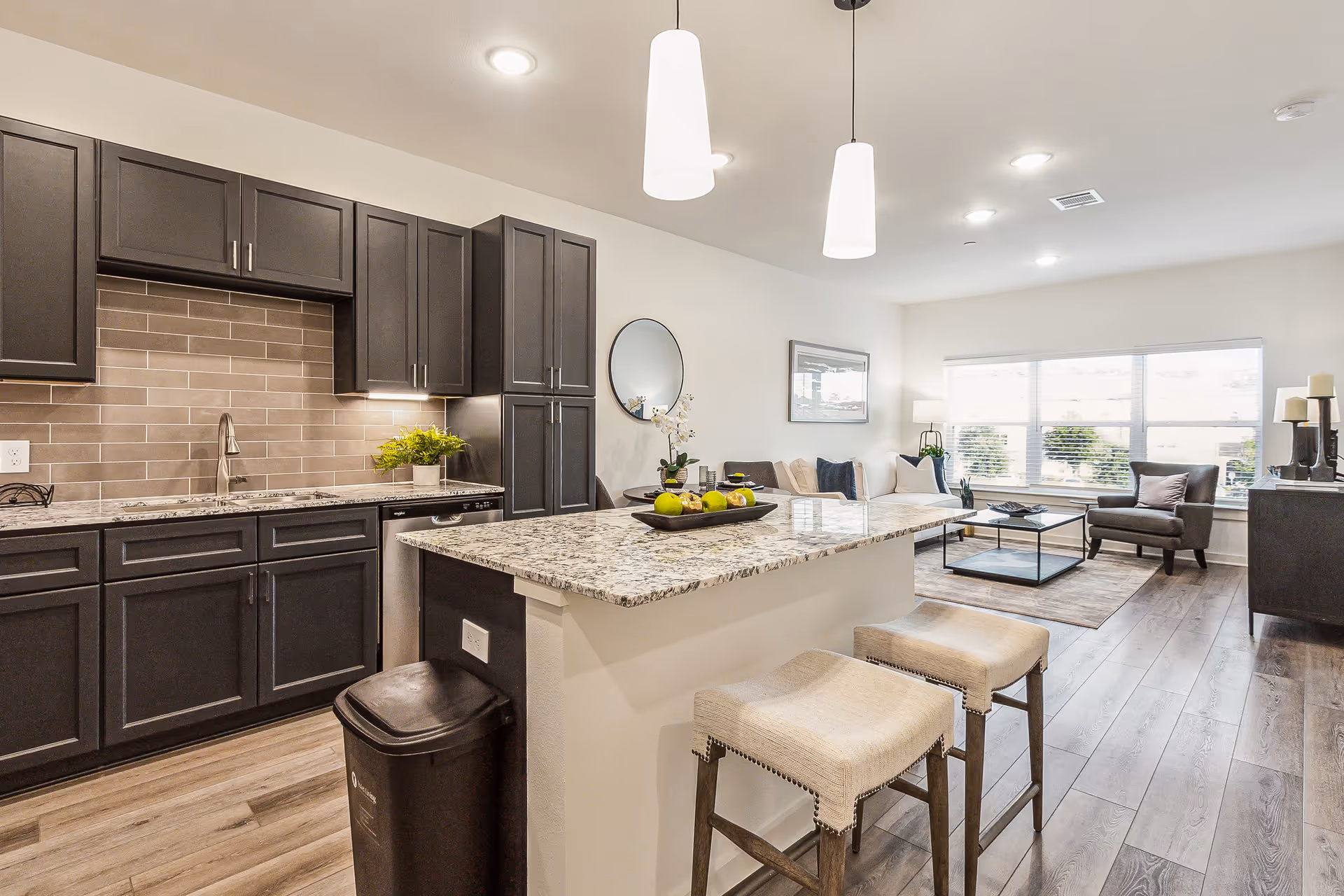 A modern open-concept kitchen and living room area with dark cabinetry, a granite countertop island with two beige cushioned stools, a trash bin, and a living room furnished with a sofa, armchair, coffee table, and large window letting in natural light.