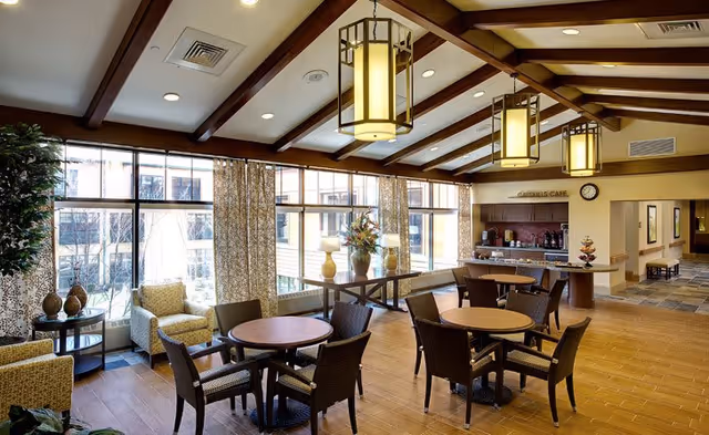 A bright and spacious common area with wooden floors and exposed wooden beams on the ceiling. The room features round tables with wicker chairs, armchairs near large windows with patterned curtains, and a counter labeled 'Cardinal's Cafe' with coffee machines and a clock on the wall. Decorative vases and plants add a welcoming touch.
