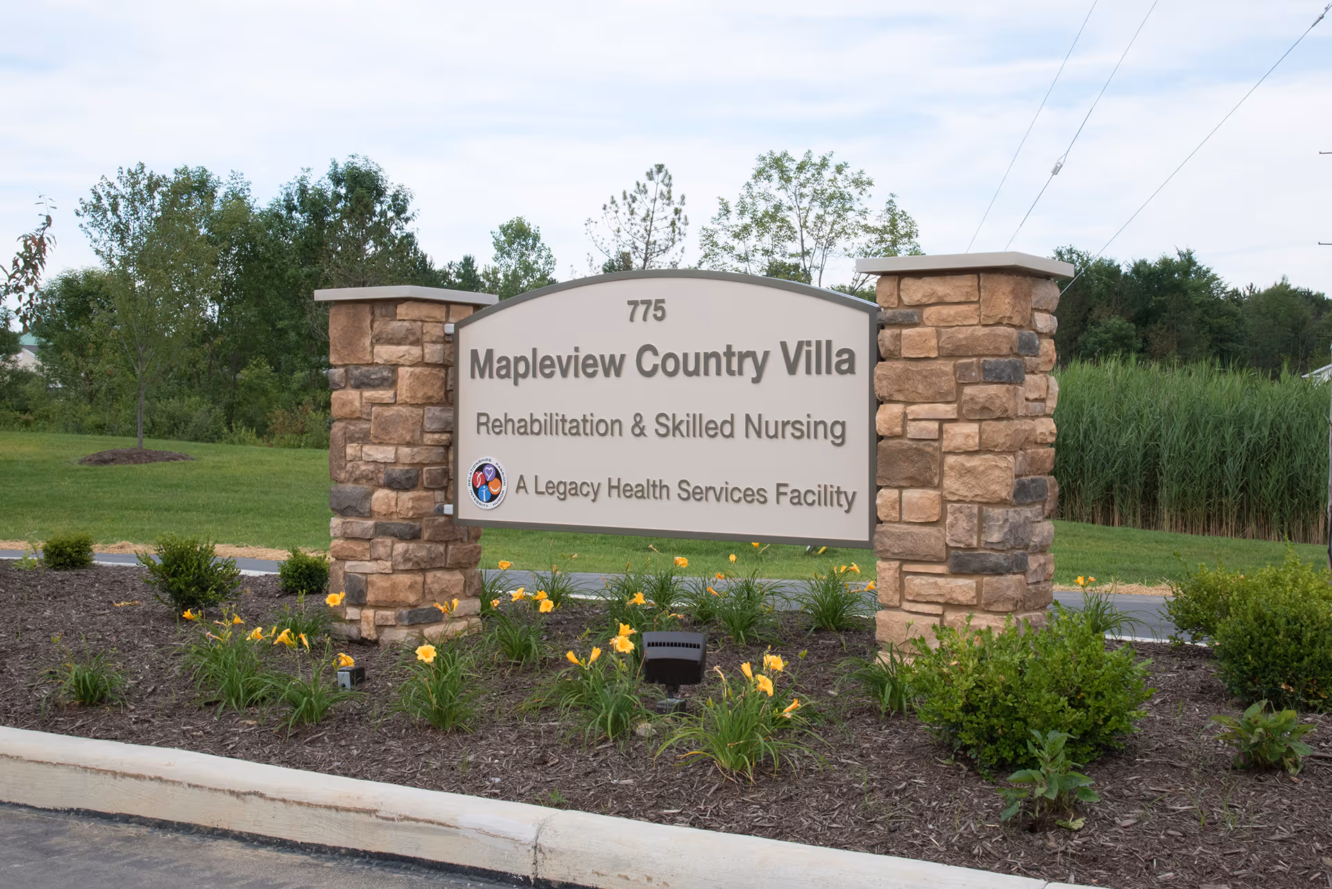Stone-pillared entrance sign reading "Mapleview Country Villa Rehabilitation & Skilled Nursing" surrounded by landscaping and flowers.
