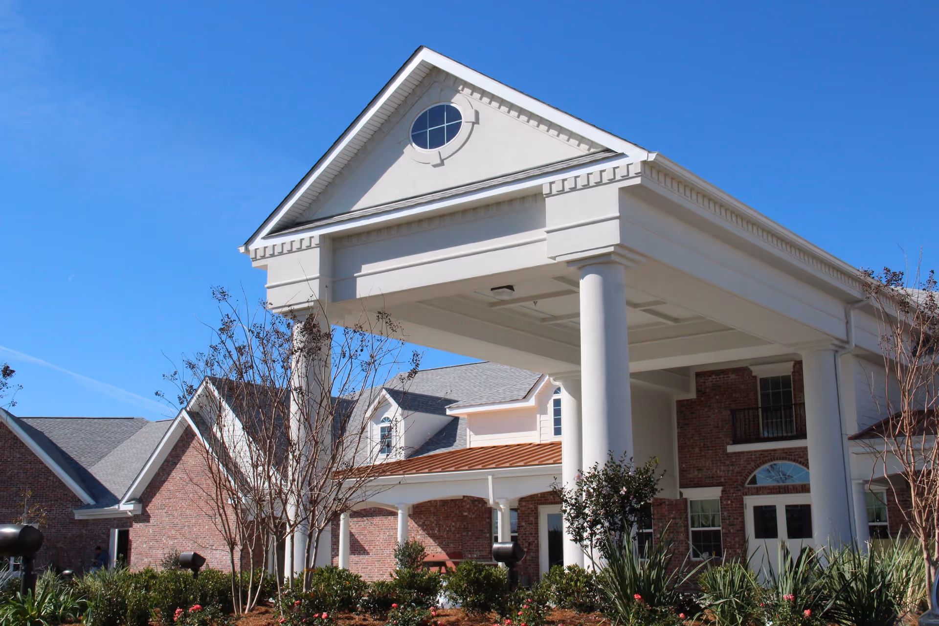 Exterior view of SummerHouse Ashton Manor featuring a large white portico with columns, brick walls, multiple windows, and a clear blue sky above. Landscaping with bushes and small trees is visible in the foreground.