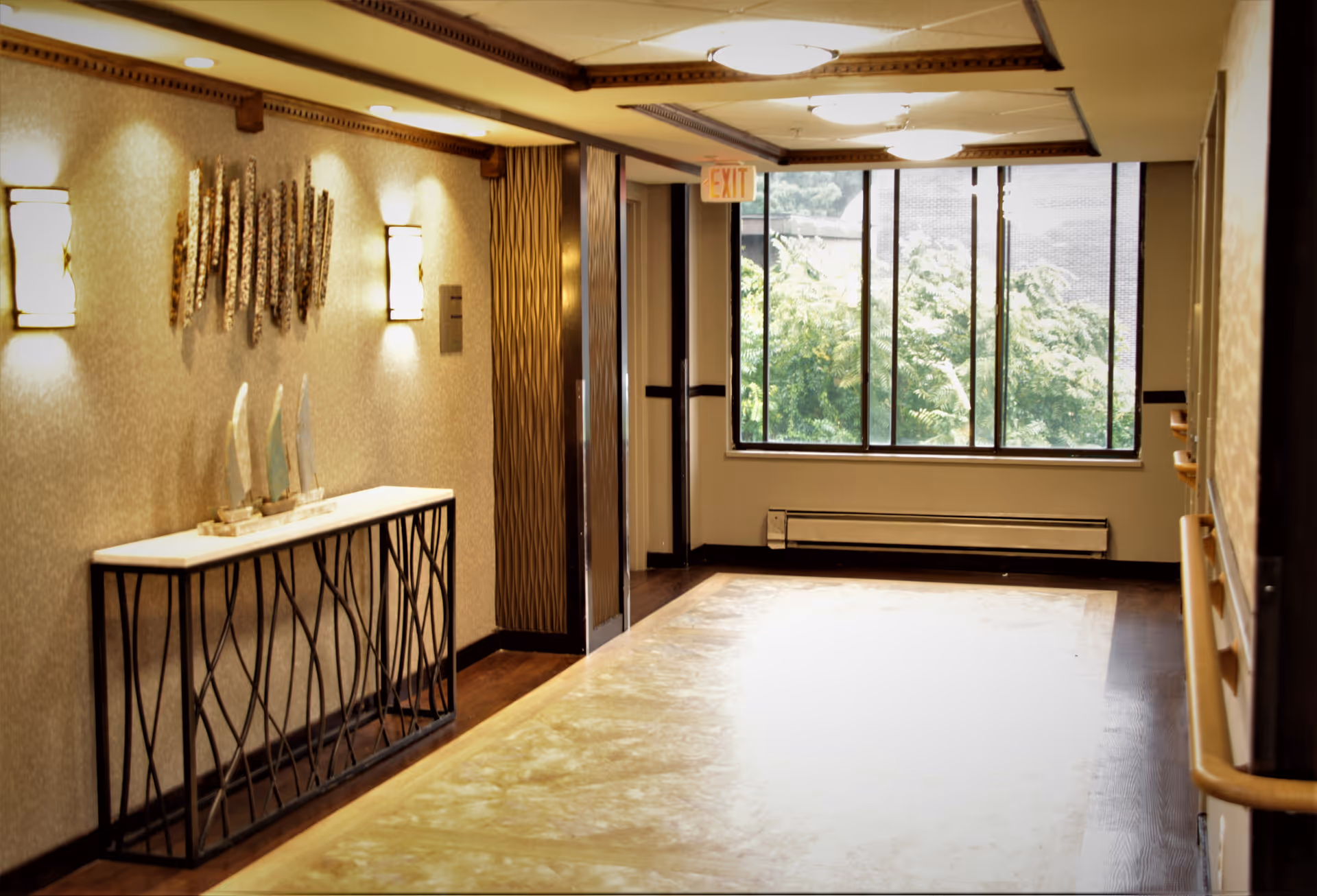Well-lit interior hallway with a decorative console table, wall sconces, handrails, and a large window showing greenery.