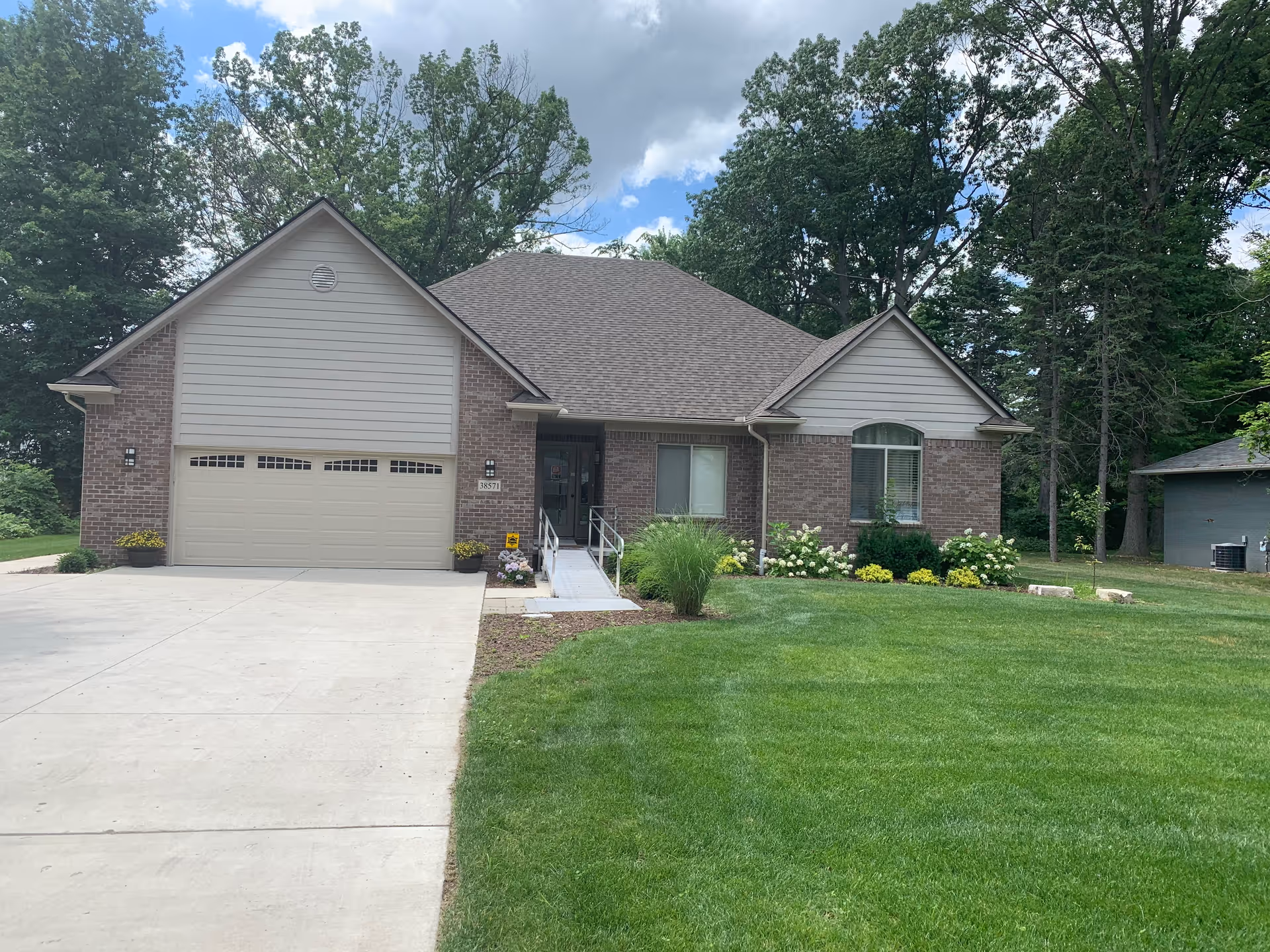 Front exterior view of a single-story brick house with a two-car garage, a concrete driveway, a small ramp leading to the front door, and a well-maintained lawn with shrubs and flowers. Trees and a partly cloudy sky are visible in the background.