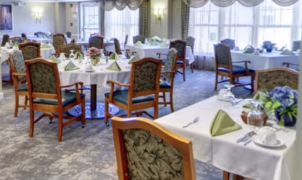 A dining room with multiple round and rectangular tables covered with white tablecloths, each set with green folded napkins, glassware, and floral centerpieces. The room has patterned carpet, upholstered wooden chairs, large windows with drapes, and soft lighting from wall sconces.