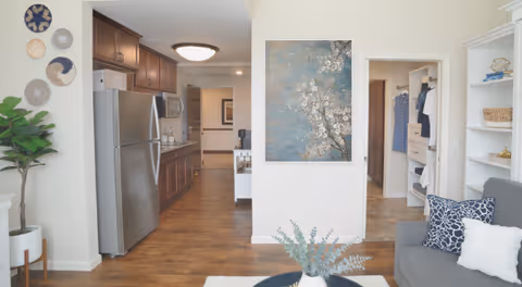 Interior view of a senior living facility showing a kitchen area with stainless steel refrigerator and wooden cabinets on the left, a living room area with a gray couch and decorative pillows on the right, a white bookshelf with decorative items, a plant in a white pot, and a large floral painting on the wall in the center.