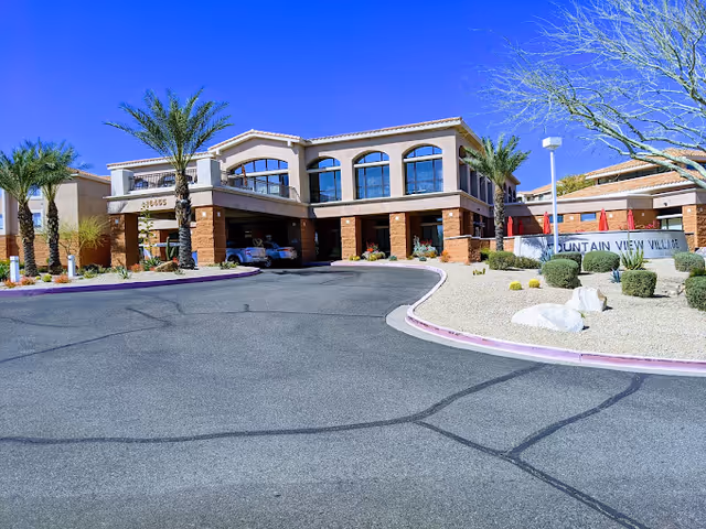 Exterior view of Fountain View Village senior living facility with a curved driveway, palm trees, desert landscaping, and a clear blue sky.