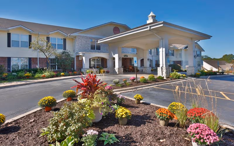 Front exterior view of Bickford of Peoria senior living facility with a covered entrance, landscaped flower beds with colorful flowers, and a clear blue sky.