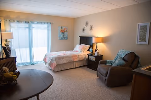 A cozy senior living bedroom featuring a single bed with a floral quilt, a bedside table with a lamp and clock, a comfortable recliner chair with a blue patterned throw, a round wooden table with a fruit basket, and large sliding glass doors with sheer curtains allowing natural light to fill the room.